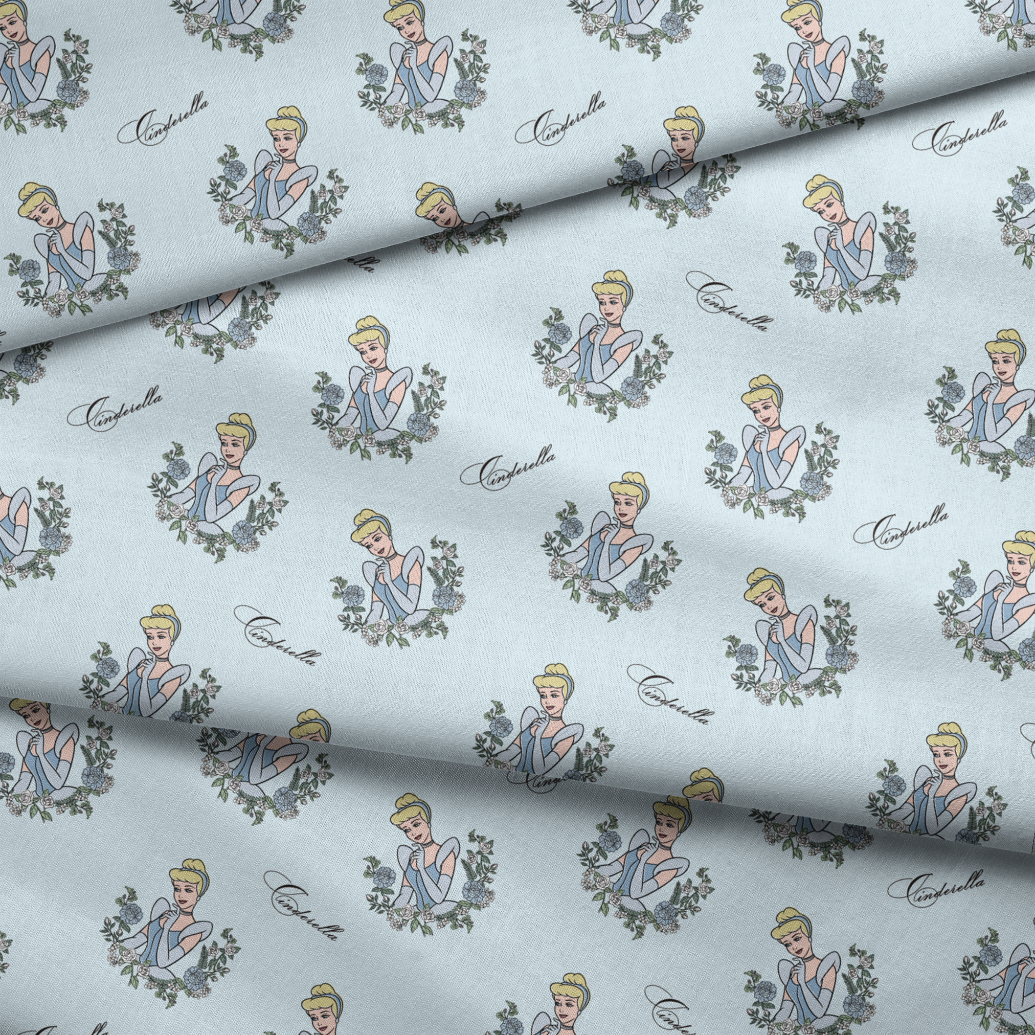 The image shows a pattern featuring a cartoon princess character in a blue dress surrounded by decorative green wreaths on a light blue background. The word "Cinderella" appears in cursive script throughout the design. fabric fold mockup