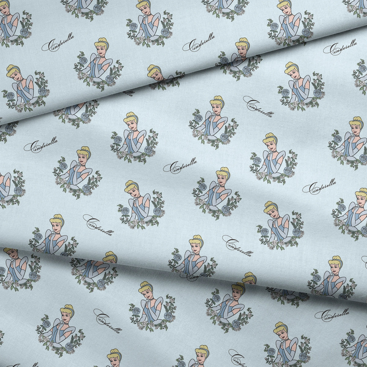 The image shows a pattern featuring a cartoon princess character in a blue dress surrounded by decorative green wreaths on a light blue background. The word "Cinderella" appears in cursive script throughout the design. fabric fold mockup