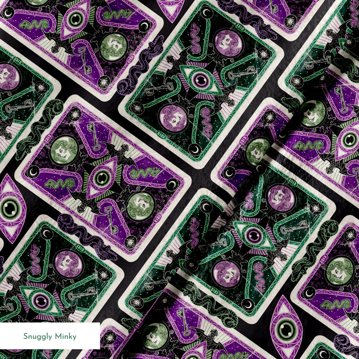Ornate playing card designs with purple and green patterns featuring central eye motifs and circular number elements against a black background. springs minky texture